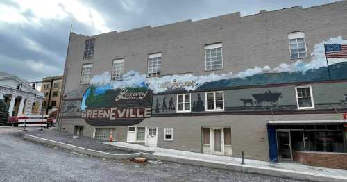 Mural on a building depicting a historical scene with "Greeneville" and a wagon, set against a cloudy sky.