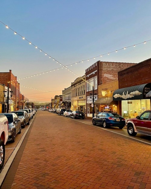 A charming street lined with shops and cars, illuminated by string lights at dusk.