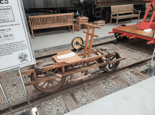 A wooden handcar on railway tracks, featuring large wheels and a simple design, displayed in a museum setting.