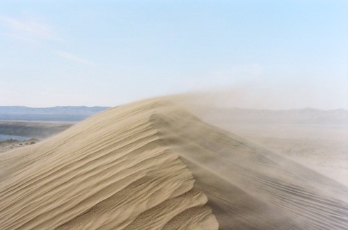 A sweeping view of a sandy dune with wind-blown sand creating ripples against a clear blue sky.