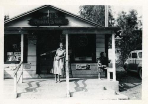 A woman stands outside a small country store, while a child plays nearby. The scene is in black and white.