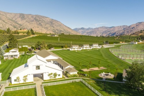 Aerial view of a scenic farm with green fields, orchards, and mountains in the background under a clear blue sky.