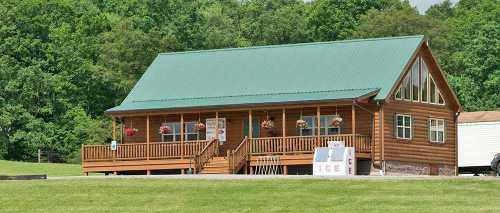 A wooden cabin with a green roof, surrounded by trees, featuring a porch and an ice sign in front.