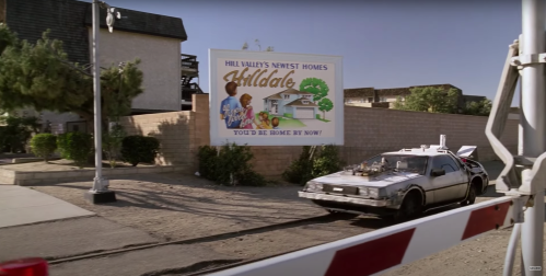 A vintage car parked near a sign for Hill Valley's newest homes, featuring a house illustration and the text "You'd be home by now!"