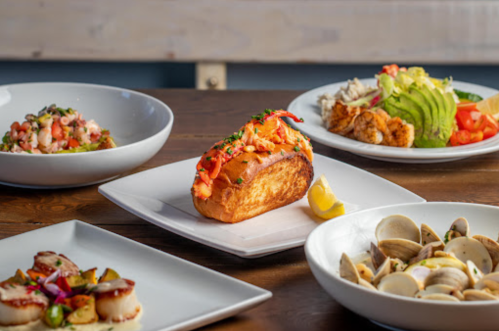 A lobster roll on a toasted bun with lemon, surrounded by various seafood dishes on white plates.
