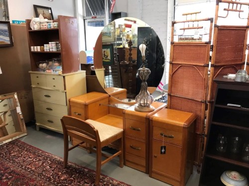 A vintage furniture display featuring a wooden vanity, mirror, chair, and various cabinets in a cozy shop setting.