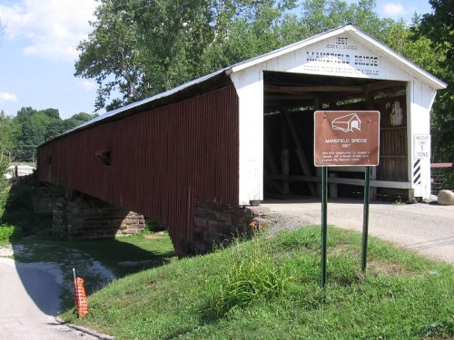 Mansfield Bridge, a historic covered bridge, with a sign and lush greenery in the background under a clear blue sky.