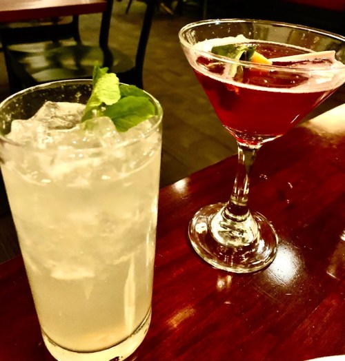A refreshing cocktail and a vibrant martini sit on a wooden table in a cozy bar setting.