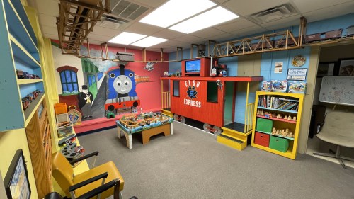 A colorful children's playroom featuring a train-themed mural, toy trains, and play areas with shelves and seating.