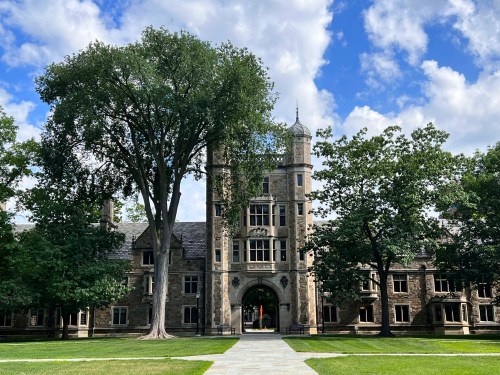 A historic stone building with tall trees and a blue sky, set in a green landscape.