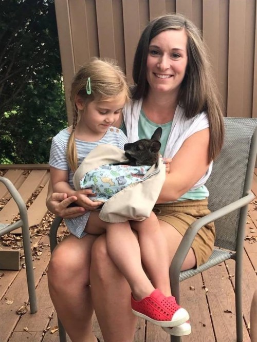 A woman and a young girl sit on a porch, with the girl holding a small dog wrapped in a blanket.