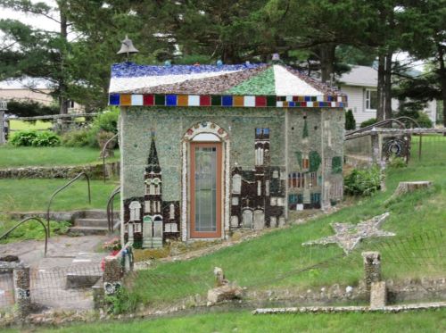 A small, colorful mosaic house surrounded by greenery and decorative elements in a garden setting.