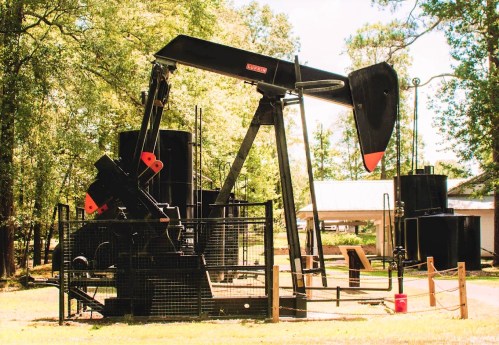 An oil pump jack in a grassy area, surrounded by trees and equipment, under a clear blue sky.
