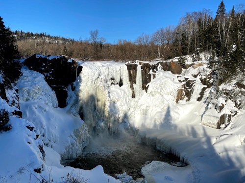 A frozen waterfall surrounded by snow-covered rocks and trees under a clear blue sky.