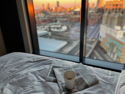 A cup of coffee on a bed with newspapers, overlooking a city skyline at sunset through a large window.