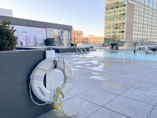 A lifebuoy hangs on a wall near a rooftop pool with a city skyline in the background.