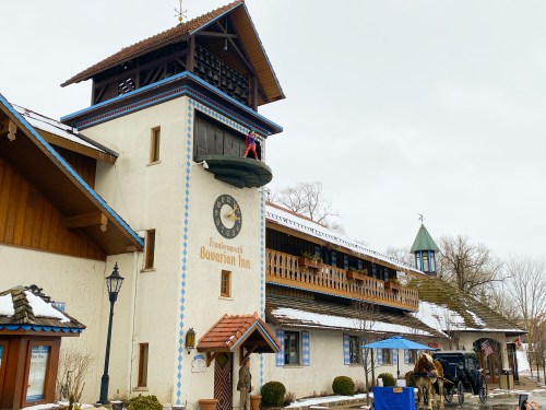 A charming Bavarian-style inn with a clock tower, snow-covered roof, and a horse-drawn carriage outside.