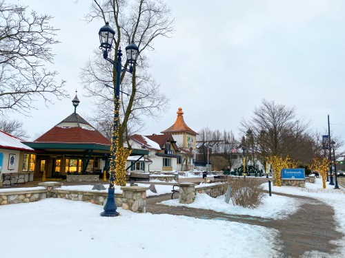 A snowy village scene with festive lights, featuring quaint buildings and a pathway lined with trees.