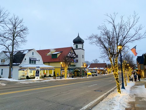 A charming winter street scene with festive lights, shops, and a tower, lined with snow-covered trees and an American flag.