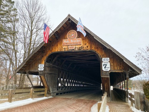 A wooden covered bridge with flags, featuring a sign that reads "Holz Brücke" and weight limit signs.
