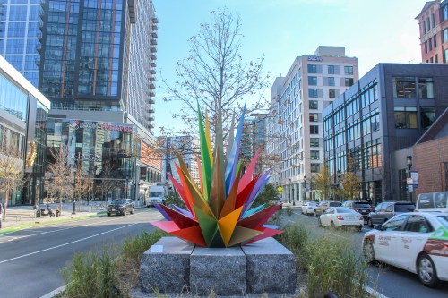 Colorful geometric sculpture surrounded by buildings and cars in an urban setting.