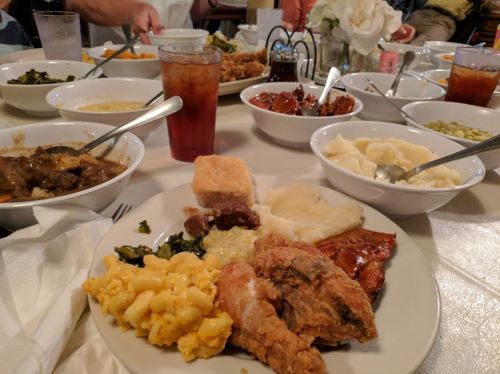 A table filled with various dishes, including fried chicken, macaroni and cheese, greens, and mashed potatoes.