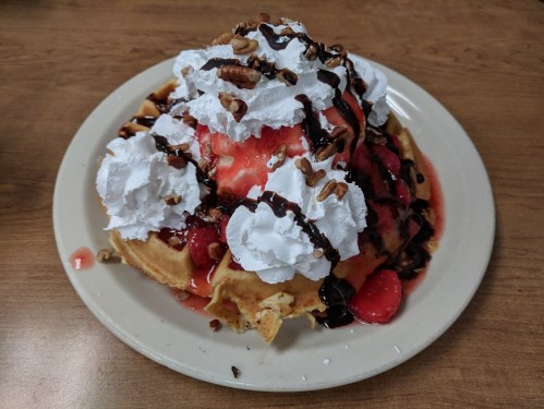 A large waffle topped with whipped cream, strawberries, chocolate syrup, and chopped nuts on a white plate.