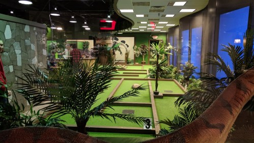 Indoor mini-golf course with lush greenery, stone walls, and a dinosaur decoration in the foreground.
