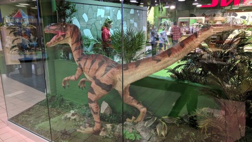 A life-sized dinosaur model stands behind glass in a vibrant indoor setting with plants and people in the background.