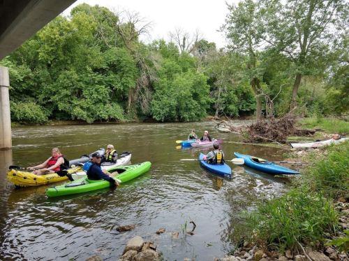 A group of kayakers in colorful kayaks paddles on a calm river surrounded by lush greenery.