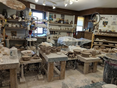 A pottery studio filled with clay pieces, tools, and shelves of finished pottery, all covered in a layer of dust.