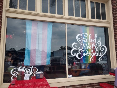 A restaurant window displays a transgender pride flag and a rainbow flag, with the text "Smoked Meat Everyday."