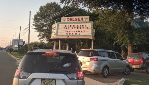 Signboard at a drive-in theater listing movies: "Lion King," "Toy Story 4," and "Yesterday." Cars parked nearby.