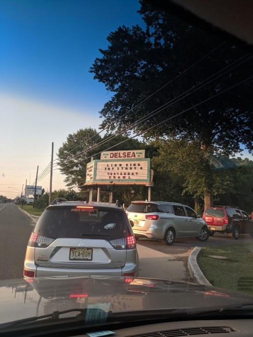 A marquee sign for Delsea with movie titles, surrounded by parked cars and trees under a clear sky.