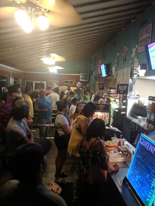 A crowded restaurant with people waiting in line to order food, featuring a menu board and various food items visible.