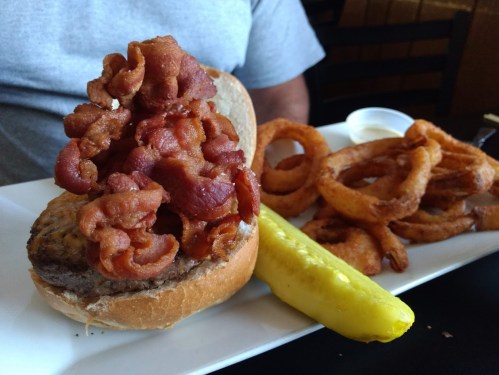 A close-up of a burger topped with crispy bacon, served with onion rings and a pickle on a white plate.