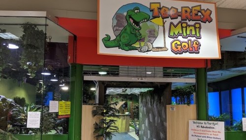 Entrance to Tee-Rex Mini Golf featuring a colorful sign and lush greenery. Fun indoor mini-golf setting.