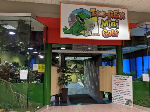 Entrance to Tee-Rex Mini Golf featuring a colorful sign, tropical decor, and a welcoming atmosphere.