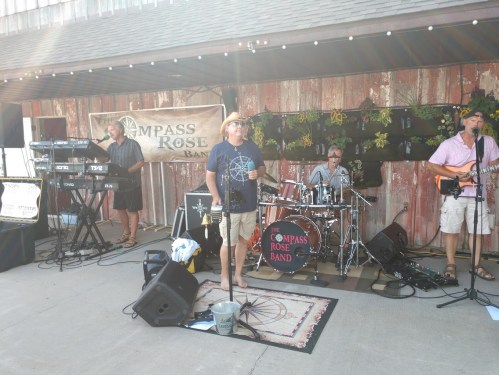 A band performs outdoors in front of a rustic backdrop, featuring a singer, drummer, and keyboardist.