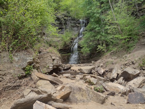 A serene waterfall cascades down rocky cliffs, surrounded by lush green trees and a rocky riverbed.