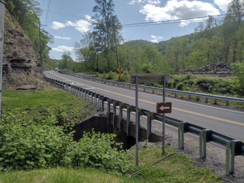 A winding road with a bridge over a small creek, surrounded by greenery and trees under a blue sky.