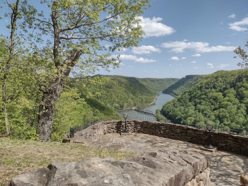 Scenic view of a river winding through green hills under a blue sky with fluffy clouds, framed by trees and stone walls.
