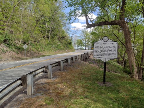 A scenic road with a historical sign for Hawks Nest Tunnel, surrounded by greenery and trees.