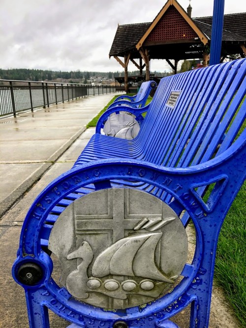 Blue bench with a decorative metal design featuring a duck and a Viking ship, overlooking a waterfront path.