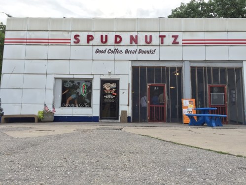 Exterior of Spudnutz, a donut shop with a sign reading "Good Coffee, Great Donuts!" and colorful outdoor seating.