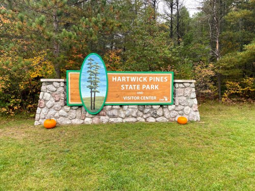 Sign for Hartwick Pines State Park and Visitor Center, surrounded by trees and pumpkins on a grassy area.