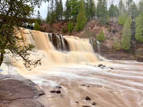 A cascading waterfall flows over rocky terrain, surrounded by lush green trees and a cloudy sky.