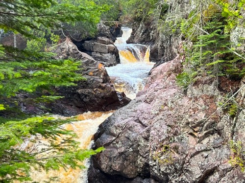 A cascading waterfall flows over rocky terrain, surrounded by lush green trees and rugged cliffs.