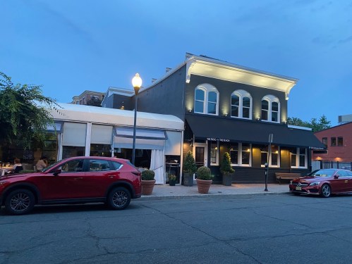 A restaurant with outdoor seating, featuring a gray building and two parked cars in front, under a twilight sky.