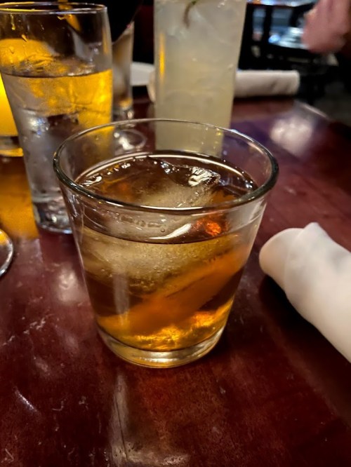 A glass of amber-colored drink with ice sits on a wooden table, surrounded by other drinks and a napkin.
